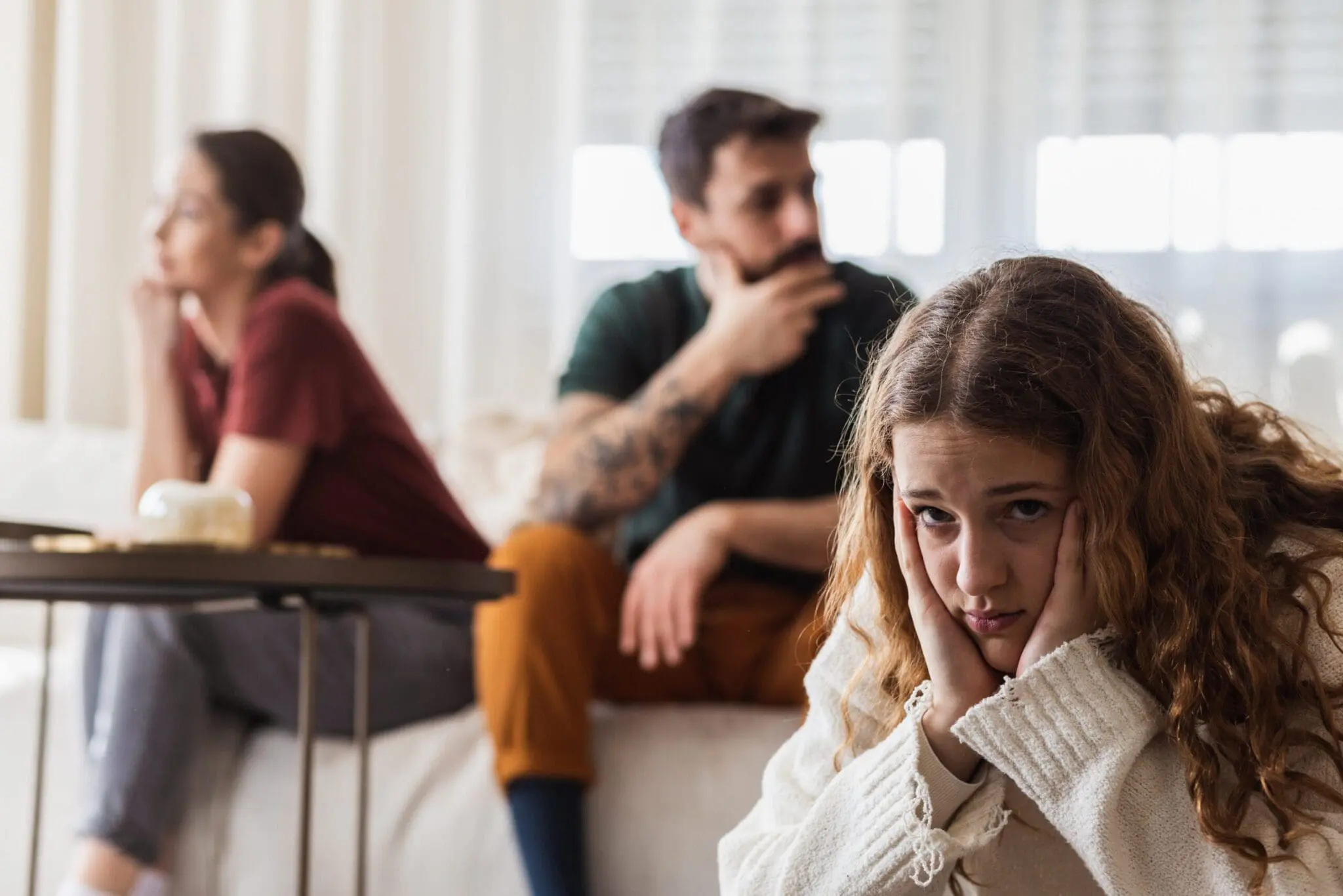 Young girl sitting in the foreground with her head resting on her hands, looking concerned, while her parents sit in the background staring away from each other. Represents family tension. Related to the Wellness blog post, 'The Need For Interpersonal Boundaries'.