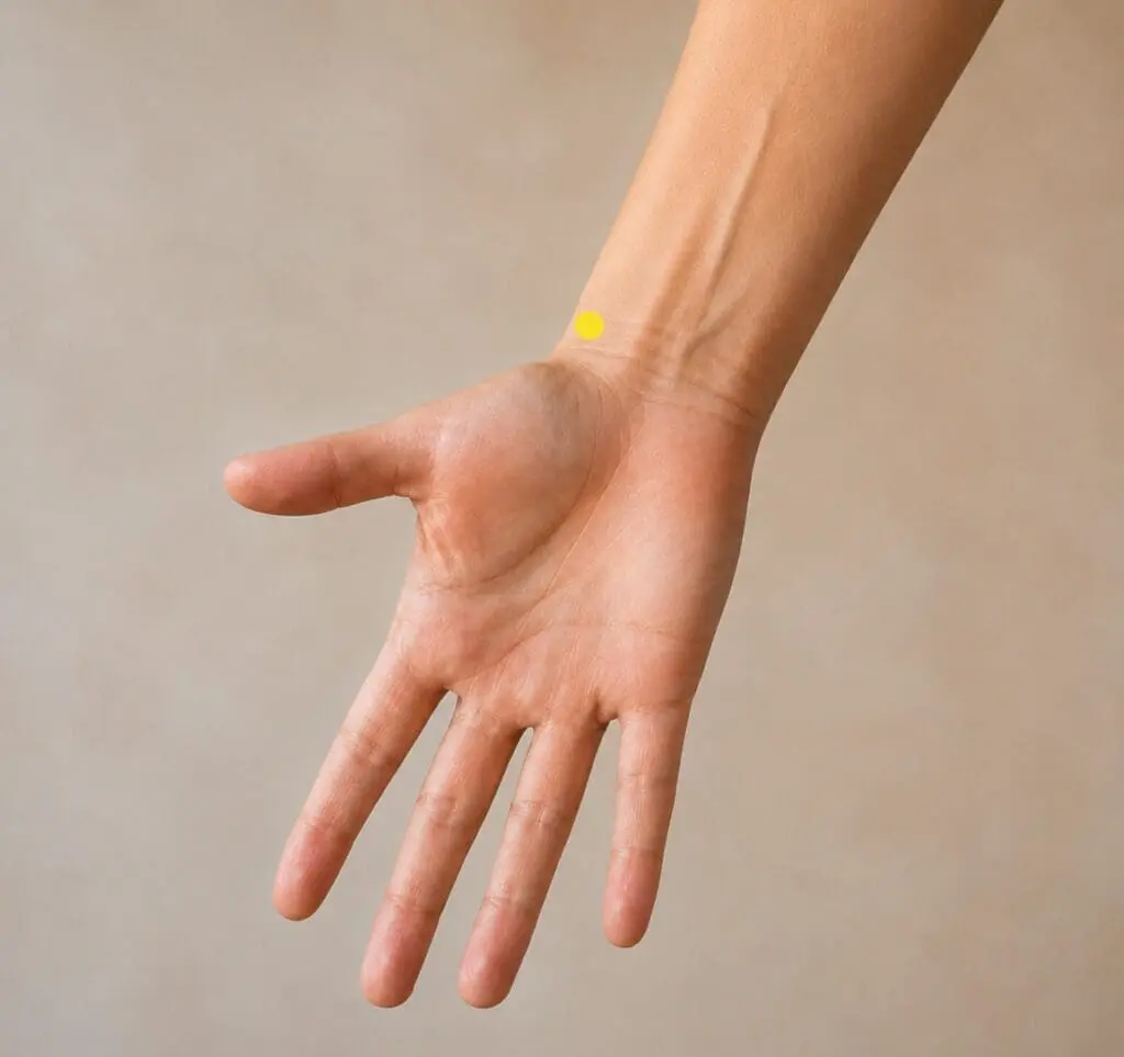 Close-up of a forearm with a yellow dot marker just above the wrist crease on the thumb side, indicating LU7.