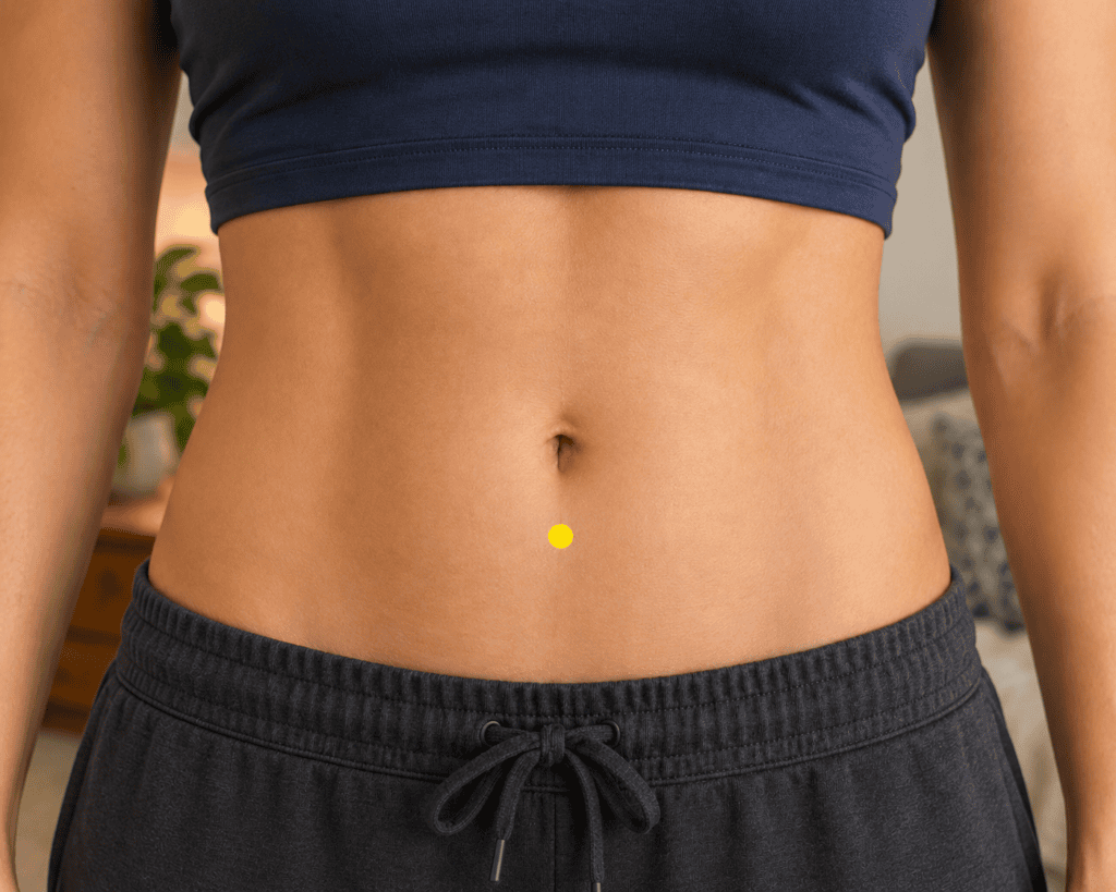 Close-up of a modestly framed lower abdomen with a yellow dot marker on the midline two finger-widths below the navel, indicating CV6.