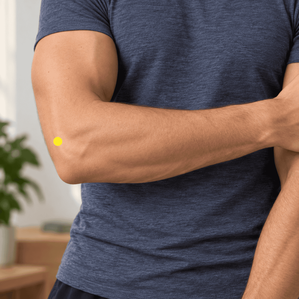 Close-up of an outer elbow with a yellow dot marker just above and outside the elbow crease near the bony bump, indicating LI12.