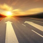 A road stretching into the distance with two arrows pointing forward, leading toward a sunset, symbolizing direction, choice, and the journey ahead. Related to the Lifestyle blog post, 'Keep Moving Forward!'