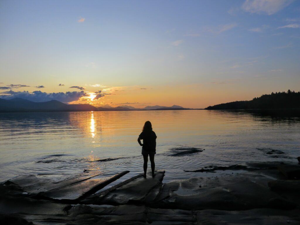 A woman standing on rocky shore, overlooking calm water at sunrise, embracing the serene and peaceful view. Related to the Lifestyle blog post, 'What Do You Desire?'