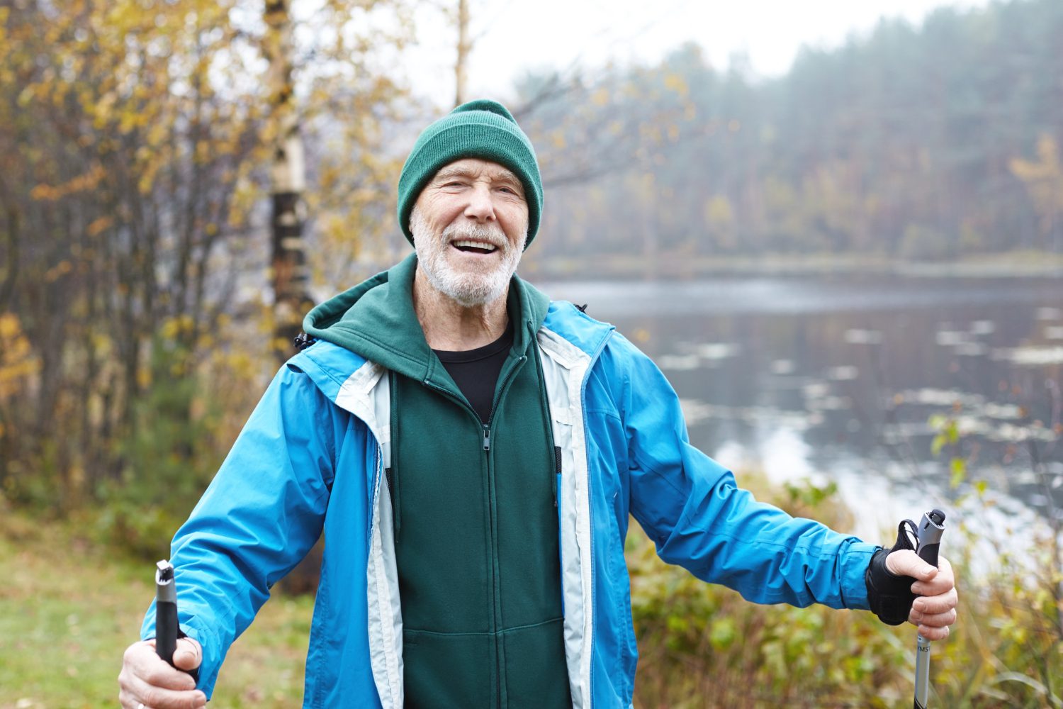 A smiling senior man with a white beard wearing a green beanie and blue jacket while using trekking poles outdoors.