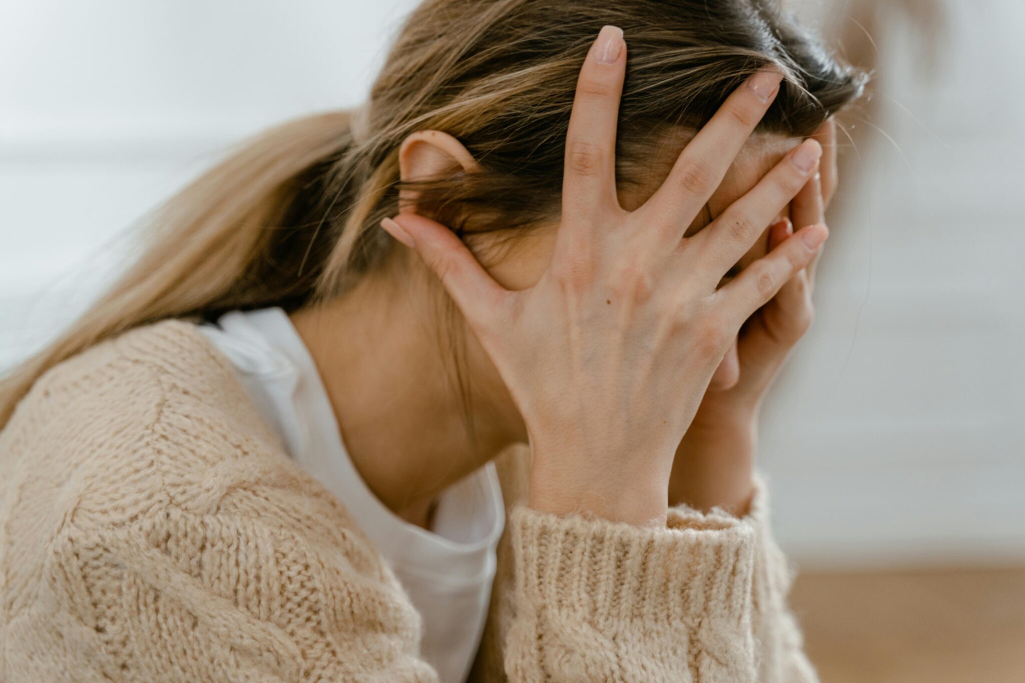 A woman in a tan sweater sitting with her face buried in her hands, illustrating the physical tension and exhaustion associated with chronic anxiety.
