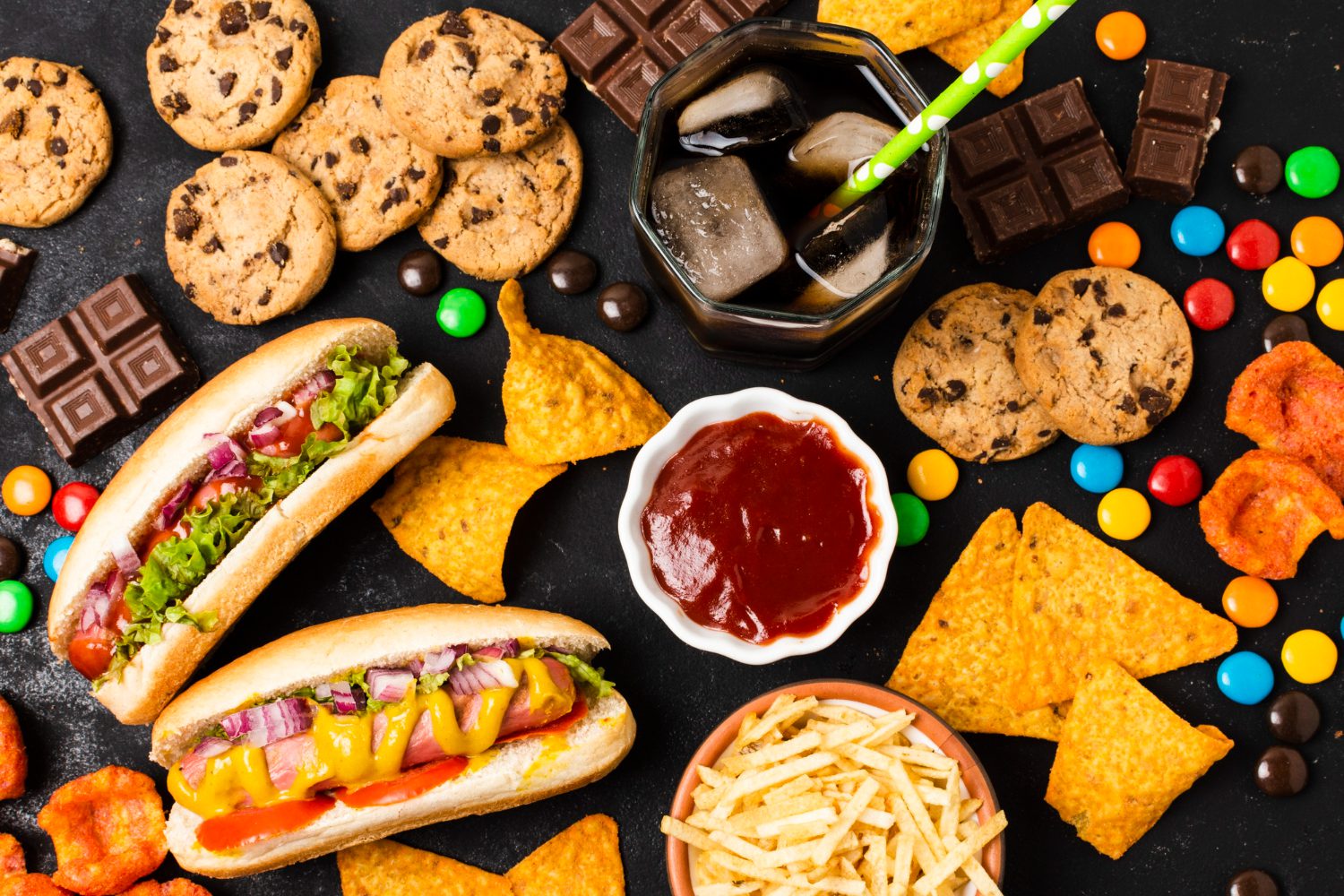 Top-down view of a spread of ultra-processed foods, including hot dogs, chocolate, cookies, and soda on a dark surface.