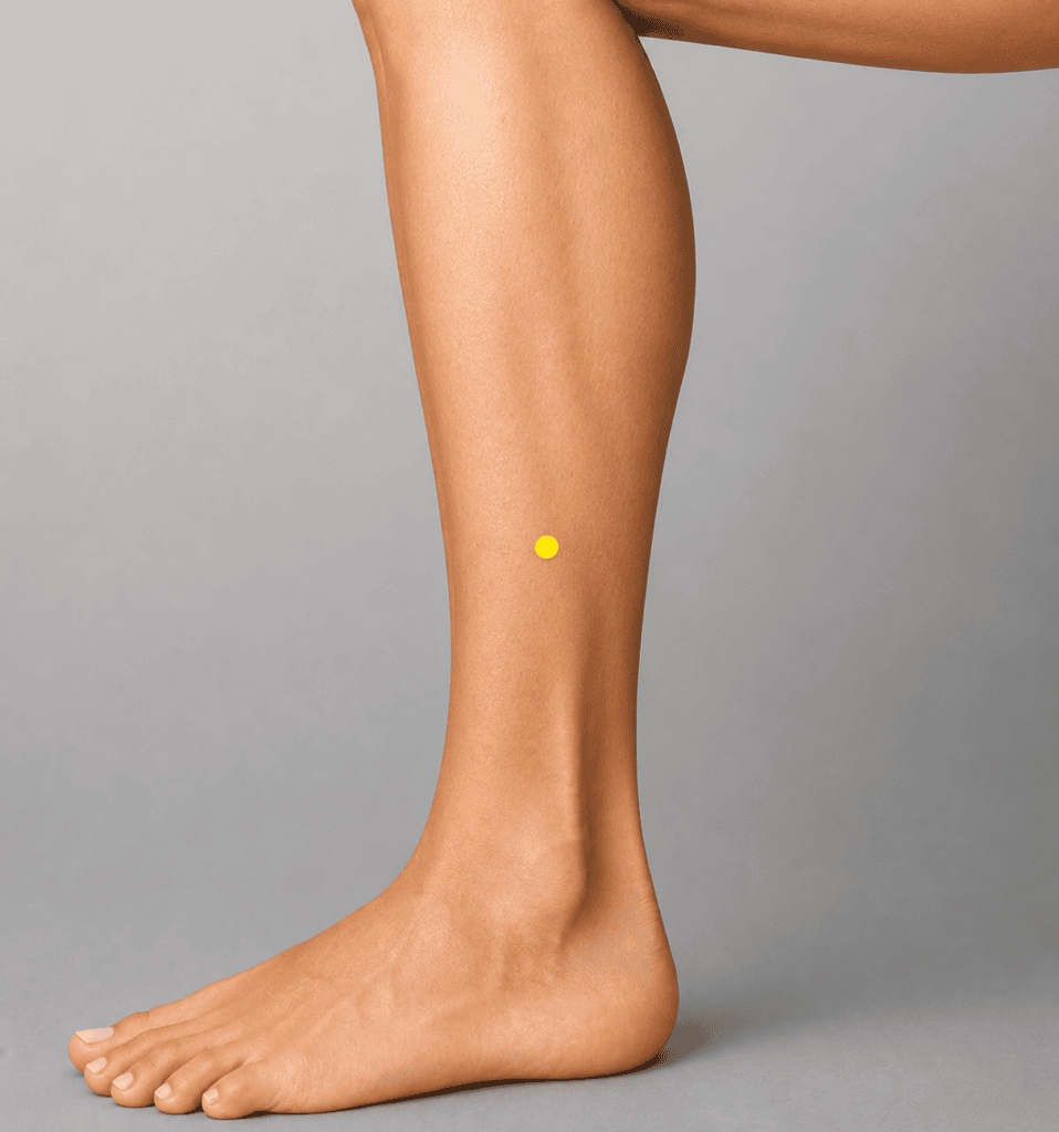 Close-up of an outer lower leg with a yellow dot marker five finger-widths above the outer ankle bone, indicating GB37.
