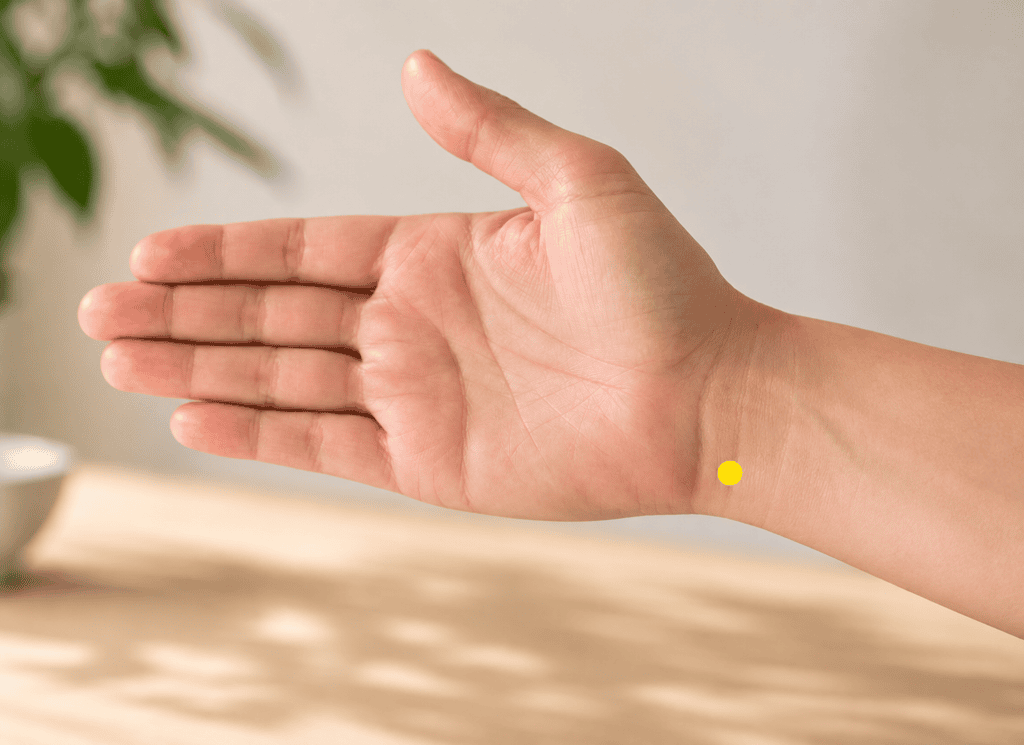 Close-up of an inner forearm with a yellow dot marker about half a thumb-width above the wrist crease on the pinky side just inside the tendon, indicating the location of HT6.