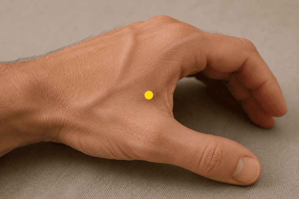 Close-up of a hand with a yellow dot marker in the soft tissue webbing between the thumb and index finger, indicating the location of LI4.