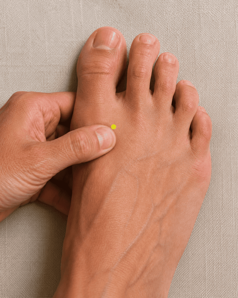 Close-up of the top of a foot with a yellow dot marker in the webbing between the first and second toes about two finger-widths back from the web, indicating the location of LR3.