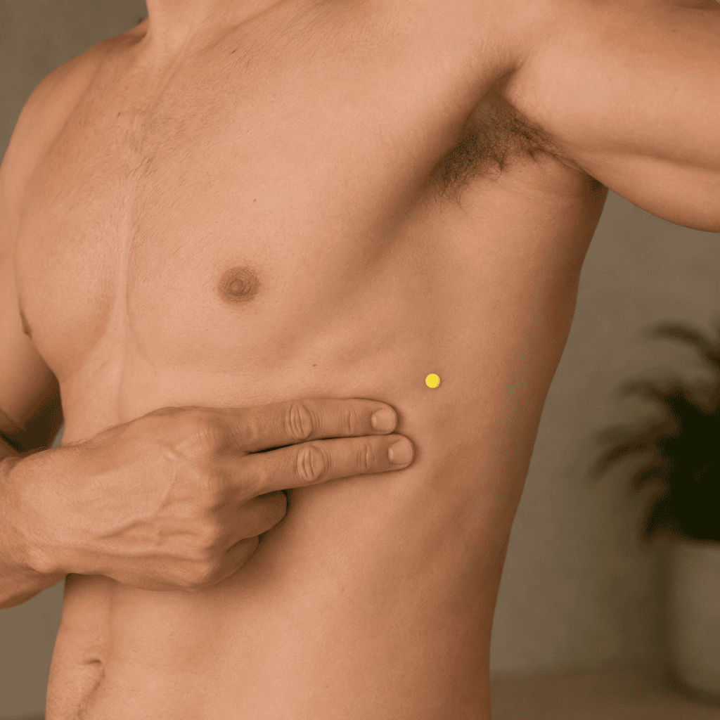 Close-up of the side of a person's ribcage with a yellow dot marker in the seventh intercostal space on the mid-axillary line roughly at bra strap level, indicating the location of SP21.