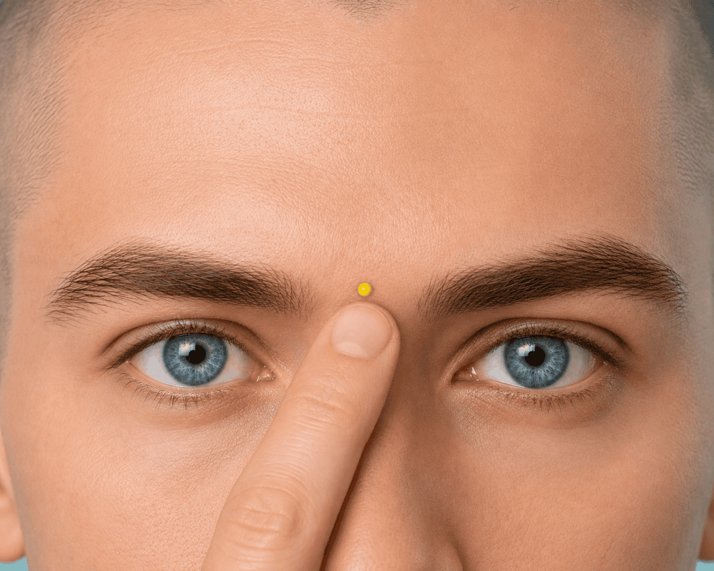 Close-up of a forehead with a yellow dot marker in the hollow between the eyebrows, indicating Yintang.