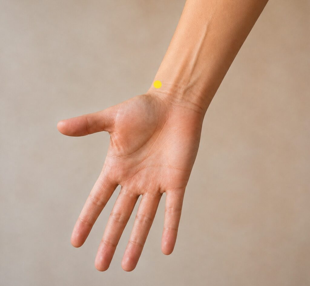 Close-up of a forearm with a yellow dot marker about two finger-widths above the wrist crease on the thumb side in a small depression on the outer edge of the radius bone, indicating the location of LU7.