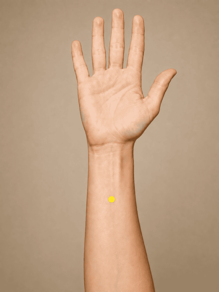 Close-up of an inner forearm with a yellow dot marker about four finger-widths above the wrist crease, one finger-width above PC6, between the two tendons, indicating the location of PC5.