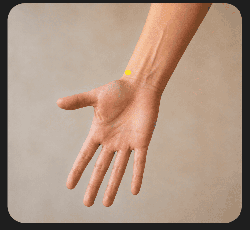 Close-up of the thumb side of an inner forearm with a yellow dot marker two finger-widths above the wrist crease, indicating LU7.