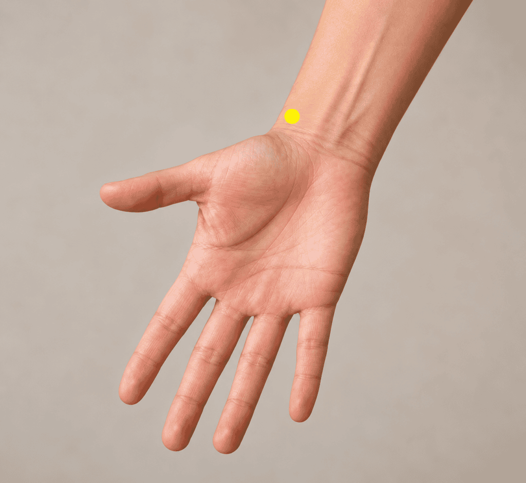 Close-up of the thumb side of an inner forearm with a yellow dot marker two finger-widths above the wrist crease, indicating LU7.